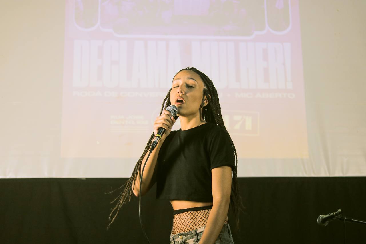 Mulher negra, com tranças e cropped preto, está em primeiro plano cantando com um microfone na mão. Atrás dela está projetado um cartaz com a frase Declama Mulher